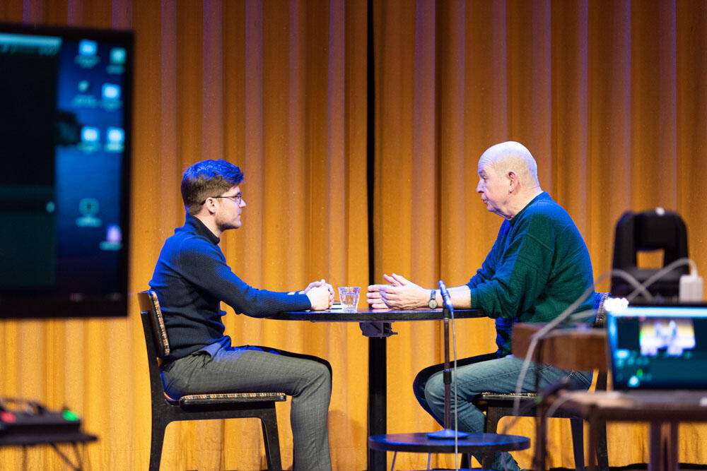 Twee mannen zitten aan een tafel, één kijkt naar de ander, op een podium met een projector op de achtergrond.