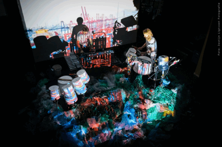 Een muzikant speelt op drums voor een kleurrijke geprojecteerde achtergrond in een donkere ruimte.