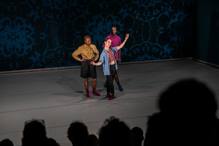 Drie performers op een podium tijdens een dansvoorstelling, met een abstracte achtergrond en publiek op de voorgrond.