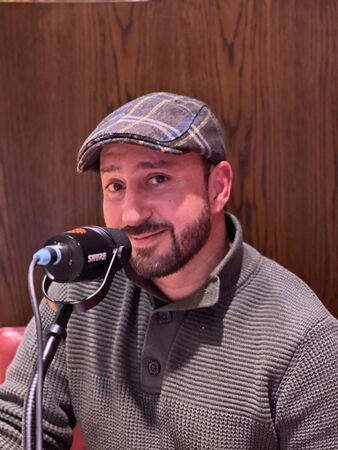 Man met een pet, zittend achter een microfoon in een warme, houten omgeving.