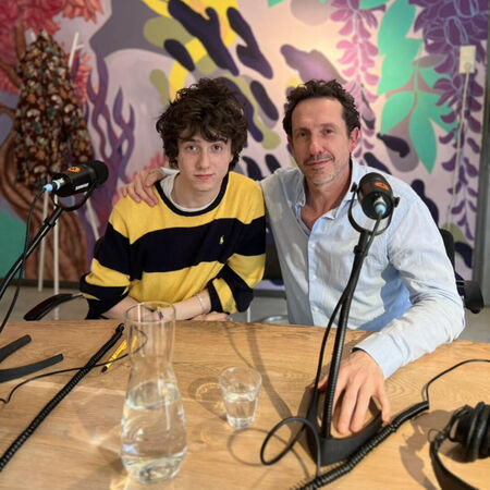 Twee mannen poseren voor een kleurrijke achtergrond met microfoons op tafel in een sfeervolle ruimte.