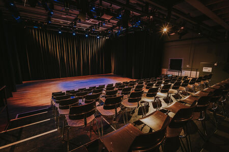 Een leeg theater met rijen stoelen en een belicht podium achter een zwarte gordijn.