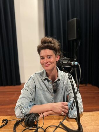 Een vrouw met een opgestoken kapsel zit aan een tafel met een microfoon en hoofdtelefoon, in een studio-achtige ruimte.