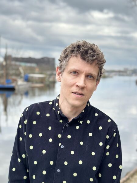 Man met gekruist haar en een shirt met stippen, staand aan een waterkant met boten op de achtergrond.