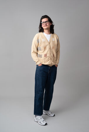 Persoon met bruin haar en een bril, gekleed in een beige cardigan en blauwe jeans, staat op een neutrale achtergrond.