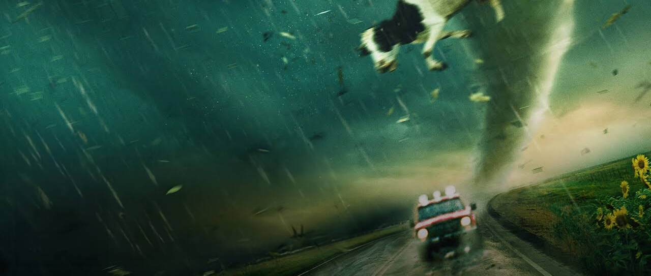 Een tornado strikt een koe en nadert een weg met een vrachtwagen. Donkere wolken en regen zijn zichtbaar.
