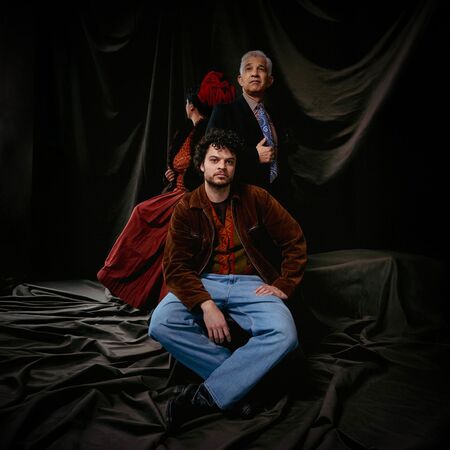 Drie personen poseren in een gedempte setting met zwarte stoffen op de achtergrond, met dramatische belichting.
