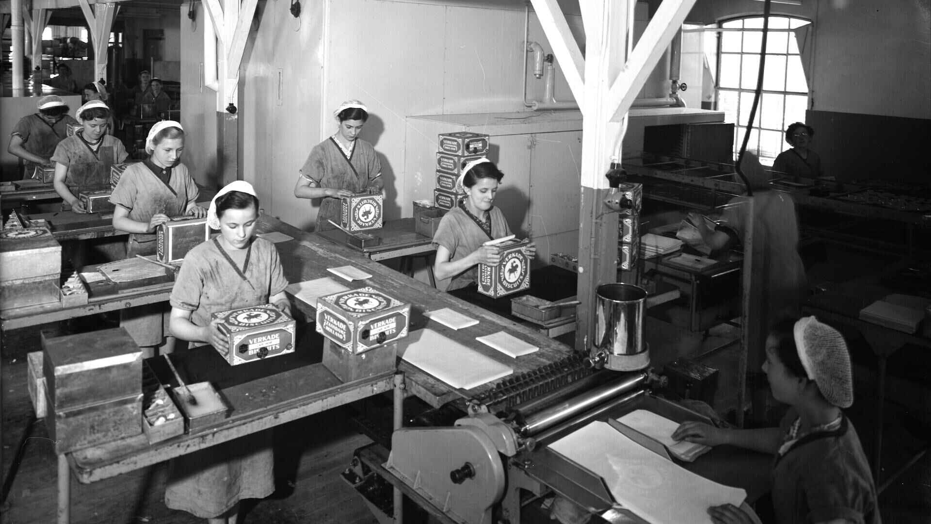 Vijf vrouwen werken aan tafels in een vintage productieomgeving, omringd door machines en materialen.