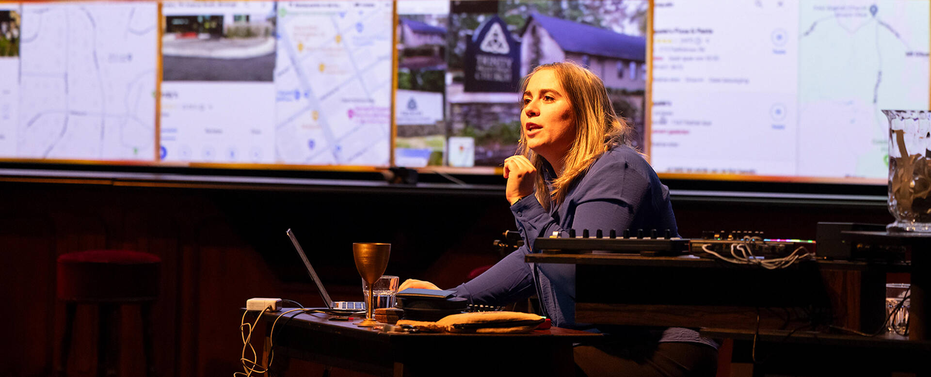 Een vrouw achter een bureau met een laptop, omringd door schermen met kaarten en afbeeldingen, terwijl ze spreekt.