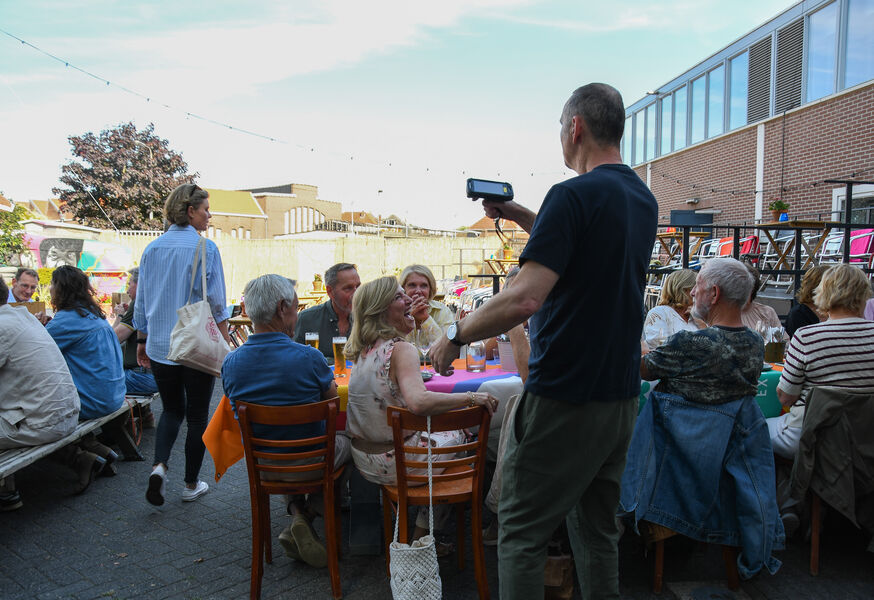 Een groep mensen zit aan een tafel in een buitenomgeving, terwijl een man met een smartphone foto’s maakt.