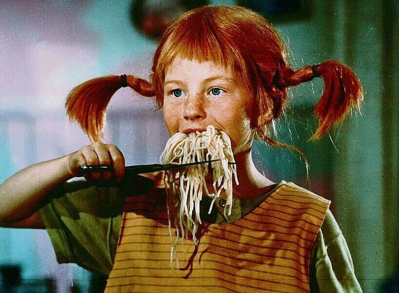 Een meisje met een oranje haardos en twee staarten houdt een schaar vast en heeft spaghetti in haar mond.