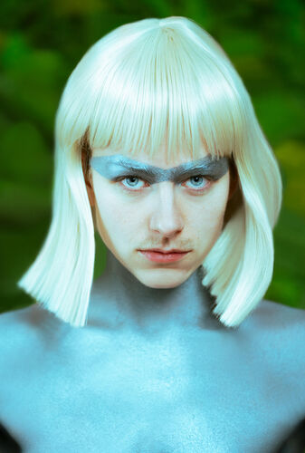 Portret van een persoon met een platinumblonde bob en blauwe gezichtsschildering, tegen een groene achtergrond.