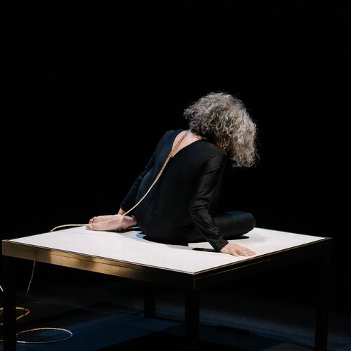 Een vrouw met krullend haar zit op een tafel, gekleed in het zwart, met een focus op haar rustige houding in een donkere omgeving.