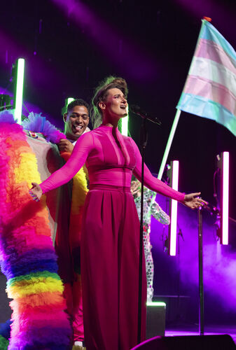 Zangeres op het podium in een felgekleurde outfit, omringd door dansers met een transgender vlag.