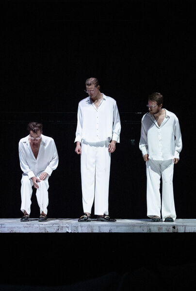 Drie mannen in witte kleding staan op een podium met een donkere achtergrond.
