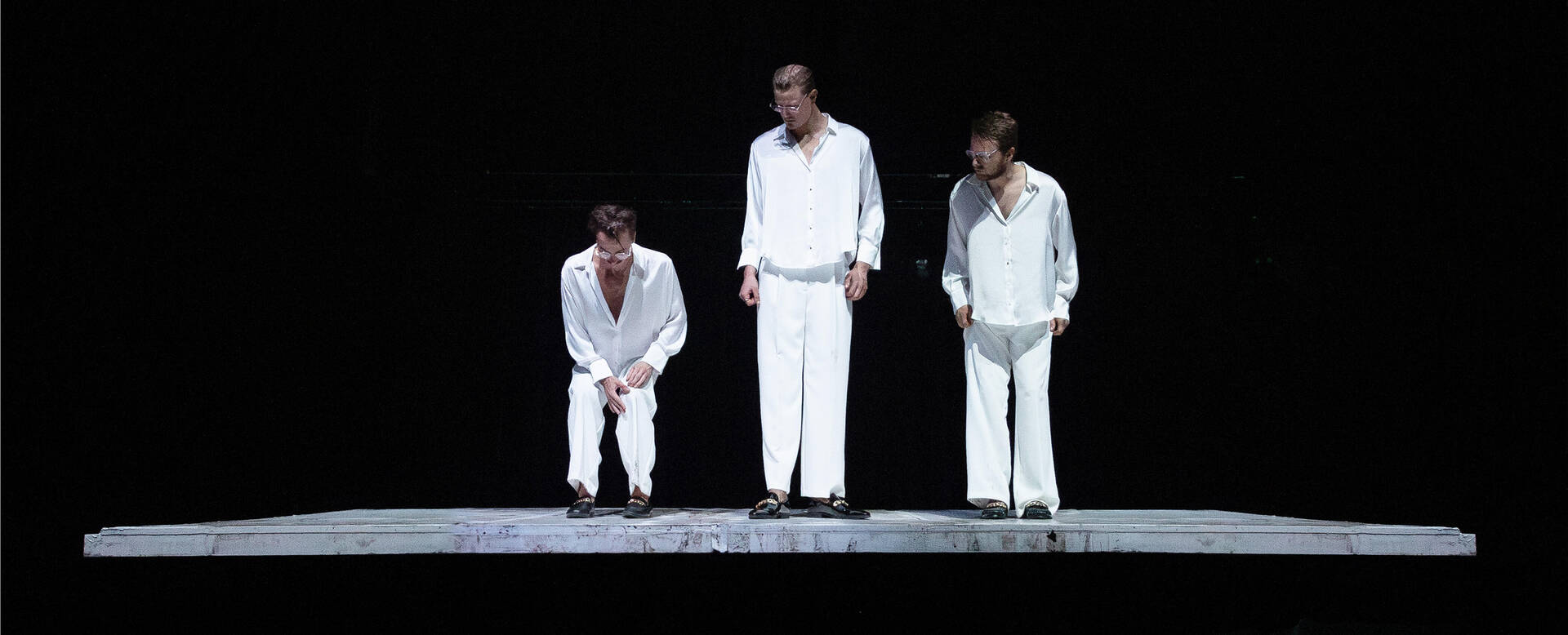 Drie mannen in witte kleding staan op een podium met zwarte achtergrond, betrokken in een dramatisch moment.