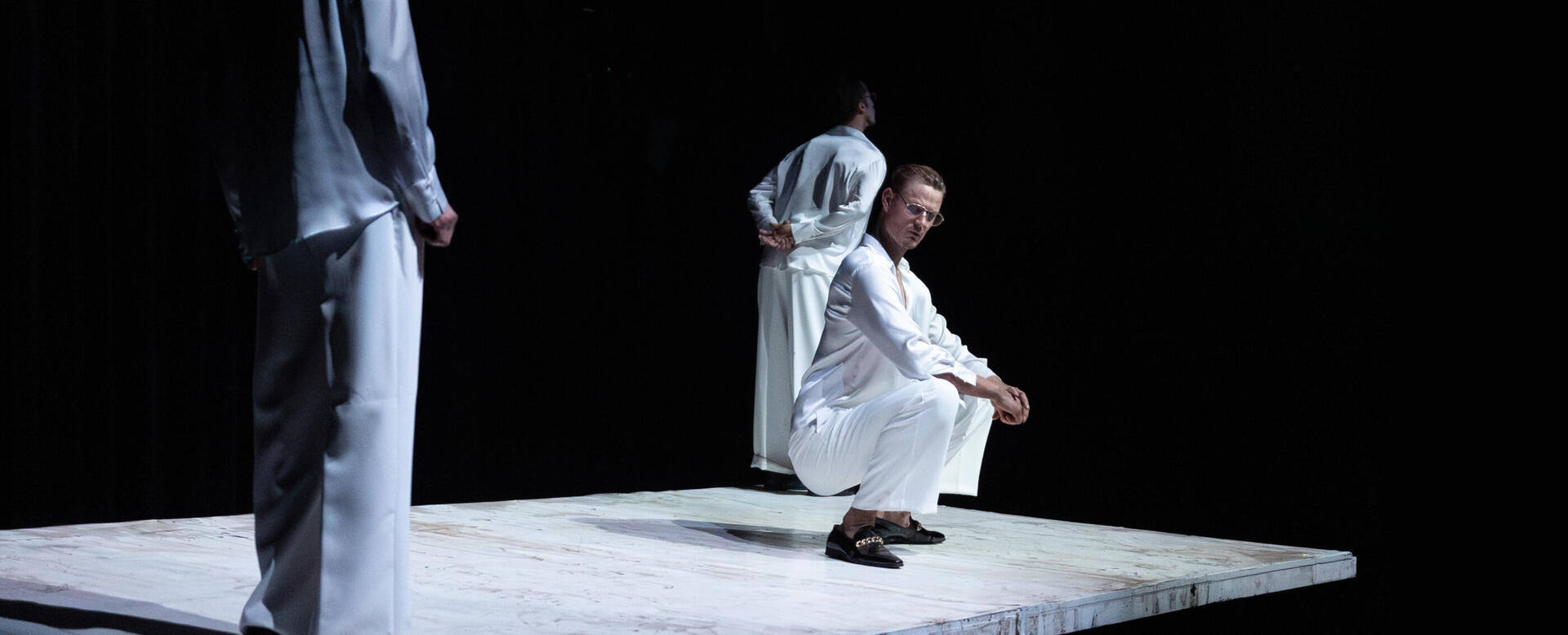 Drie mensen in witte kleding staan en zitten op een lichte, abstracte vloer in een donkere omgeving.