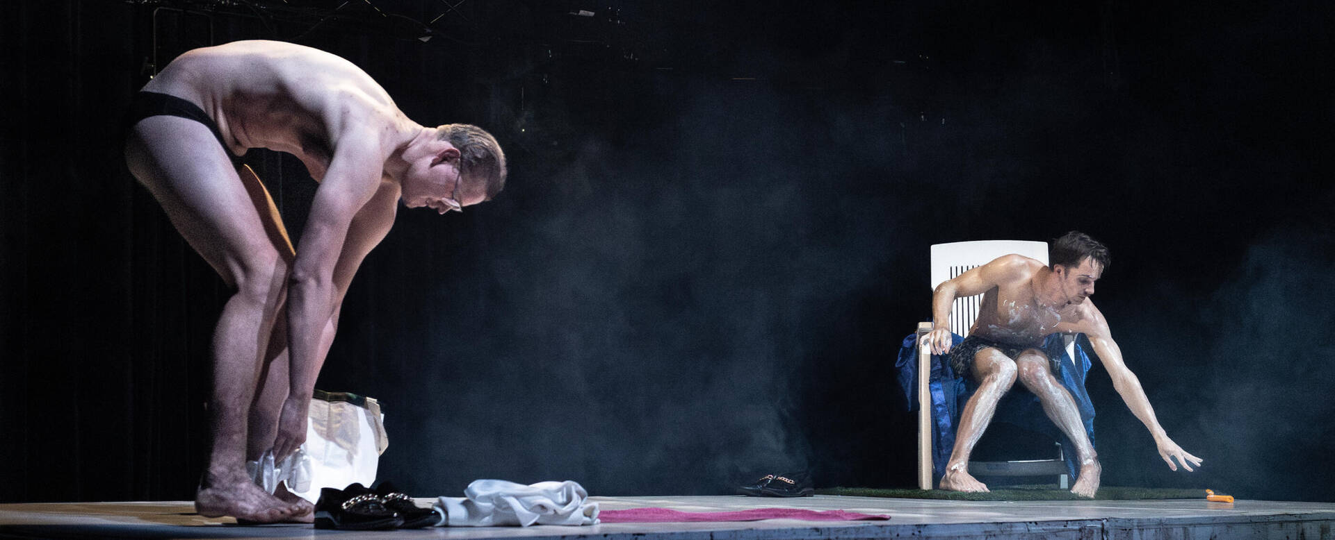 Twee naakte mannen op een podium; één bukt terwijl de ander op een stoel zit en zich vooroverbuigt. Rook om hen heen.