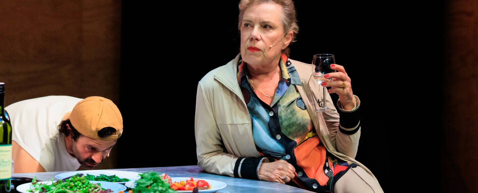 Toneelstuk met een vrouw die met een glas in de hand zit en een man die zich voorover buigt aan een tafel.