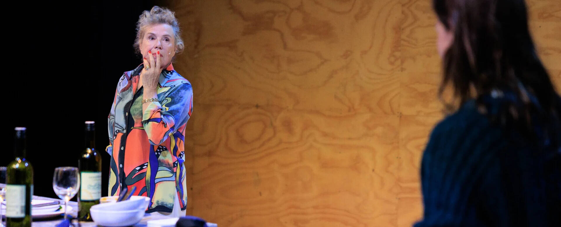 Twee acteurs op een podium, met focus op de vrouw in een kleurrijke outfit en een tafel met wijnflessen en glazen.
