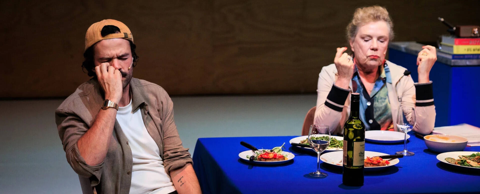 Twee personen aan een eettafel, zichtbaar onvoldaan, omringd door borden met eten en een fles wijn.