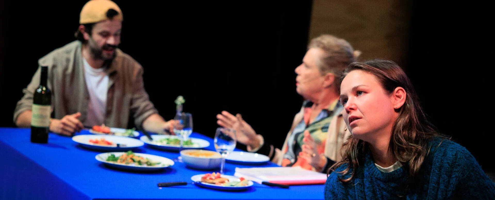 Twee vrouwen en een man aan tafel met eten en drinken, terwijl één vrouw bezorgd lijkt.