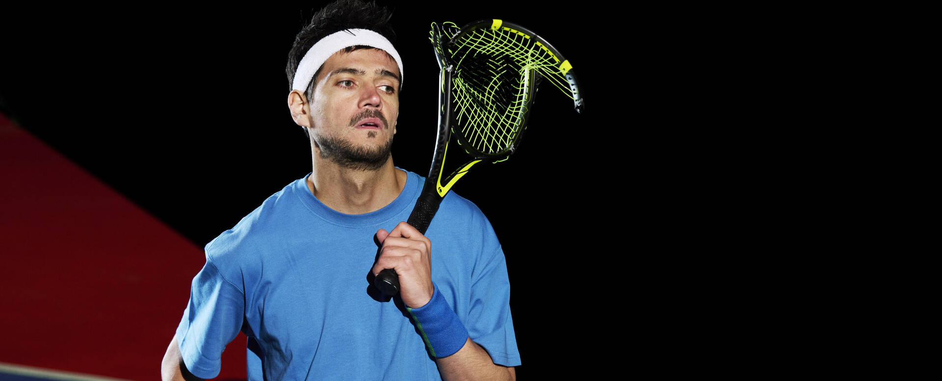 Tennisspeler in blauwe outfit houdt een racket vast, met een dynamische houding tegen een donkere achtergrond.
