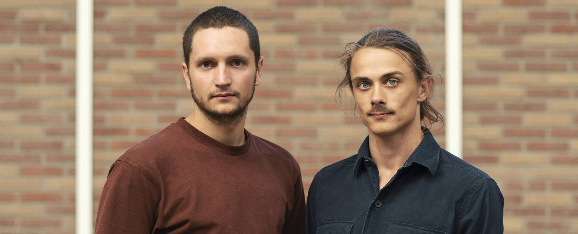 Twee mannen, één met een baard en de ander met een snor, staan naast elkaar voor een stenen muur.