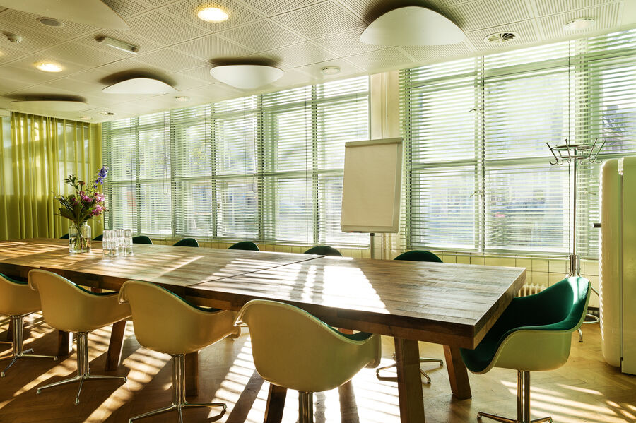 Lichte vergaderruimte met lange houten tafel, stoelen en grote ramen met uitzicht op buiten.