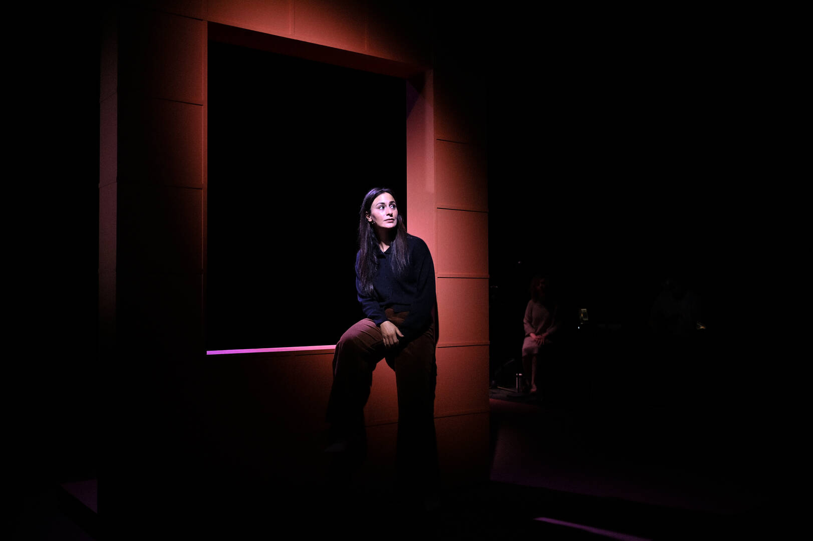 Vrouw in zwart kleding zittend in een verlichte deuropening, omgeven door schaduw.