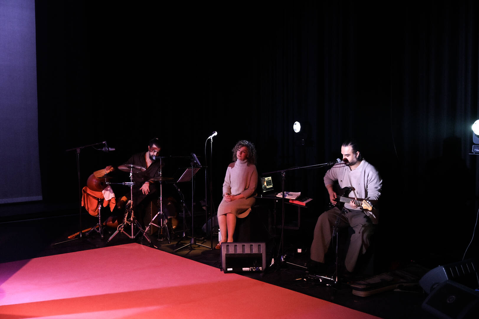 Drie muzikanten op een podium, twee met instrumenten en één op een stoel, in een donkere omgeving met lichtaccenten.