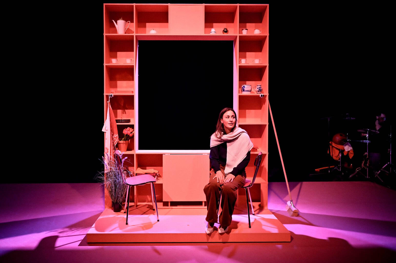 Theaterproductie met een vrouw zittend op het podium, omringd door een decor van planken en stoelen.