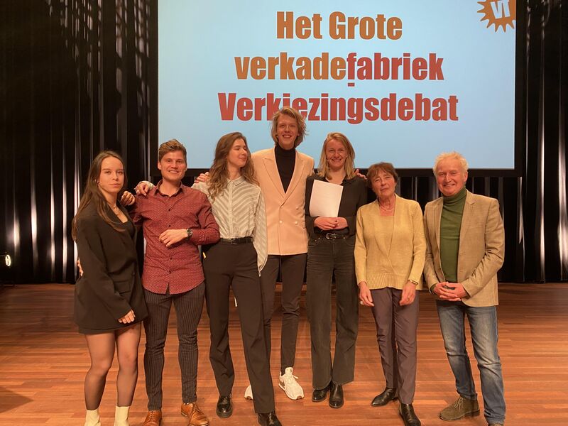 Groep mensen op het podium bij een verkiezingsdebat, met een groot scherm op de achtergrond.