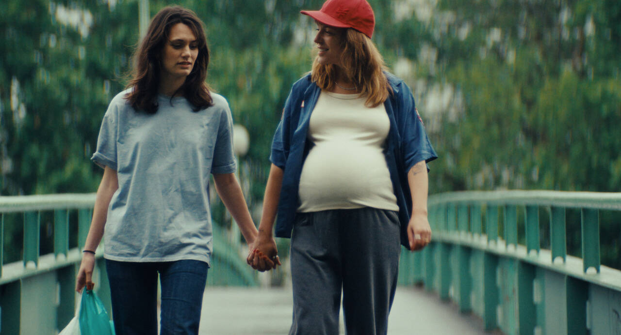 Twee vrouwen, waarvan één zichtbaar zwanger, hand in hand wandelend over een brug in een groene omgeving.