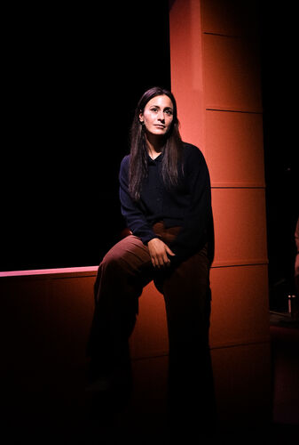 Een vrouw met lange haren zit op een rand in een donkere ruimte, met een paarse lichteffect op haar.