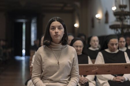 Een vrouw in een beige trui zit vooraan in een kerk, met andere vrouwen in zwarte kleding op de achtergrond.