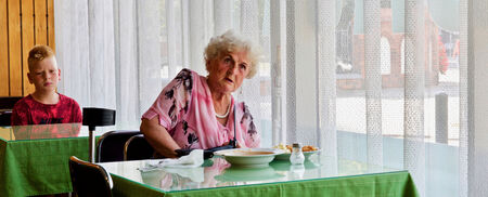 Een oudere vrouw zit aan een tafel in een restaurant, met een bord soep voor zich, terwijl een kind op de achtergrond kijkt.