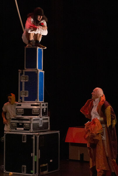 Een performer staat op een hoge stapel koffers, terwijl een andere performer beneden toekijkt in theatrale kostuums.