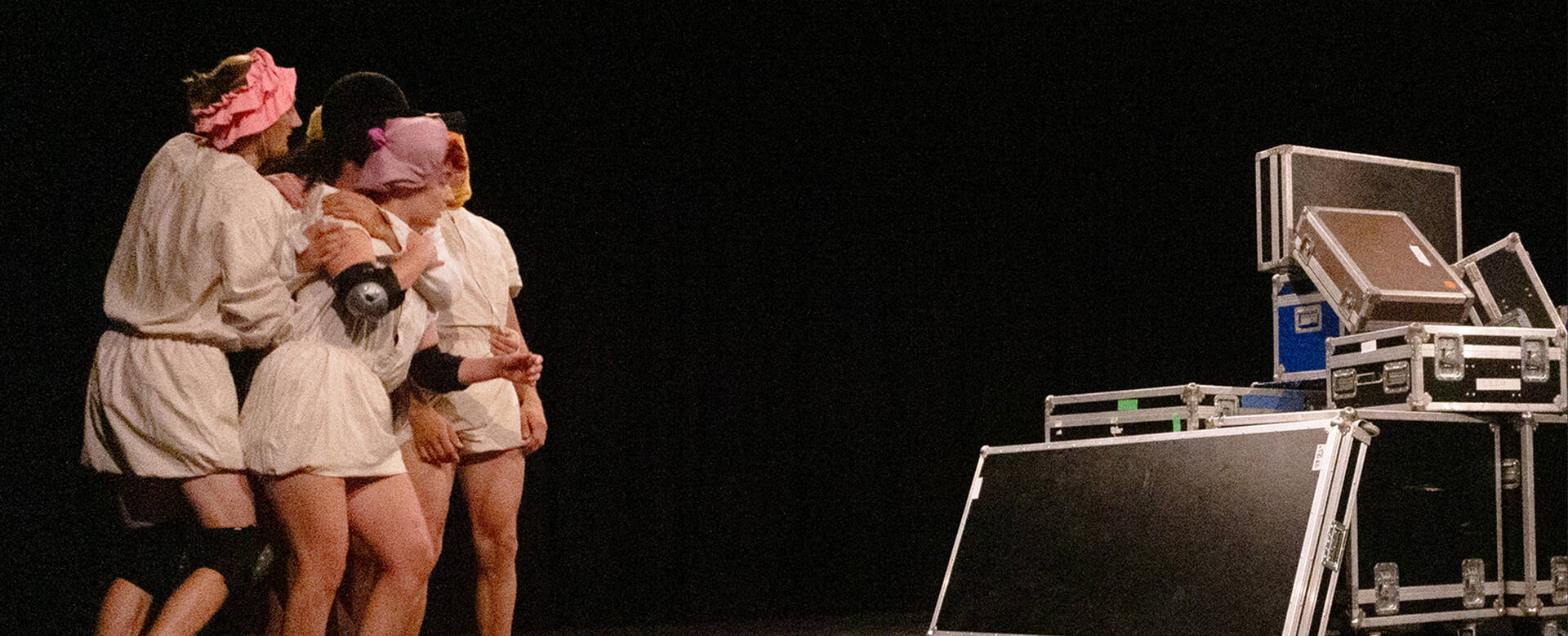 Drie personen in kostuums op het podium, omringd door apparatuur, tijdens een creatieve uitvoering.