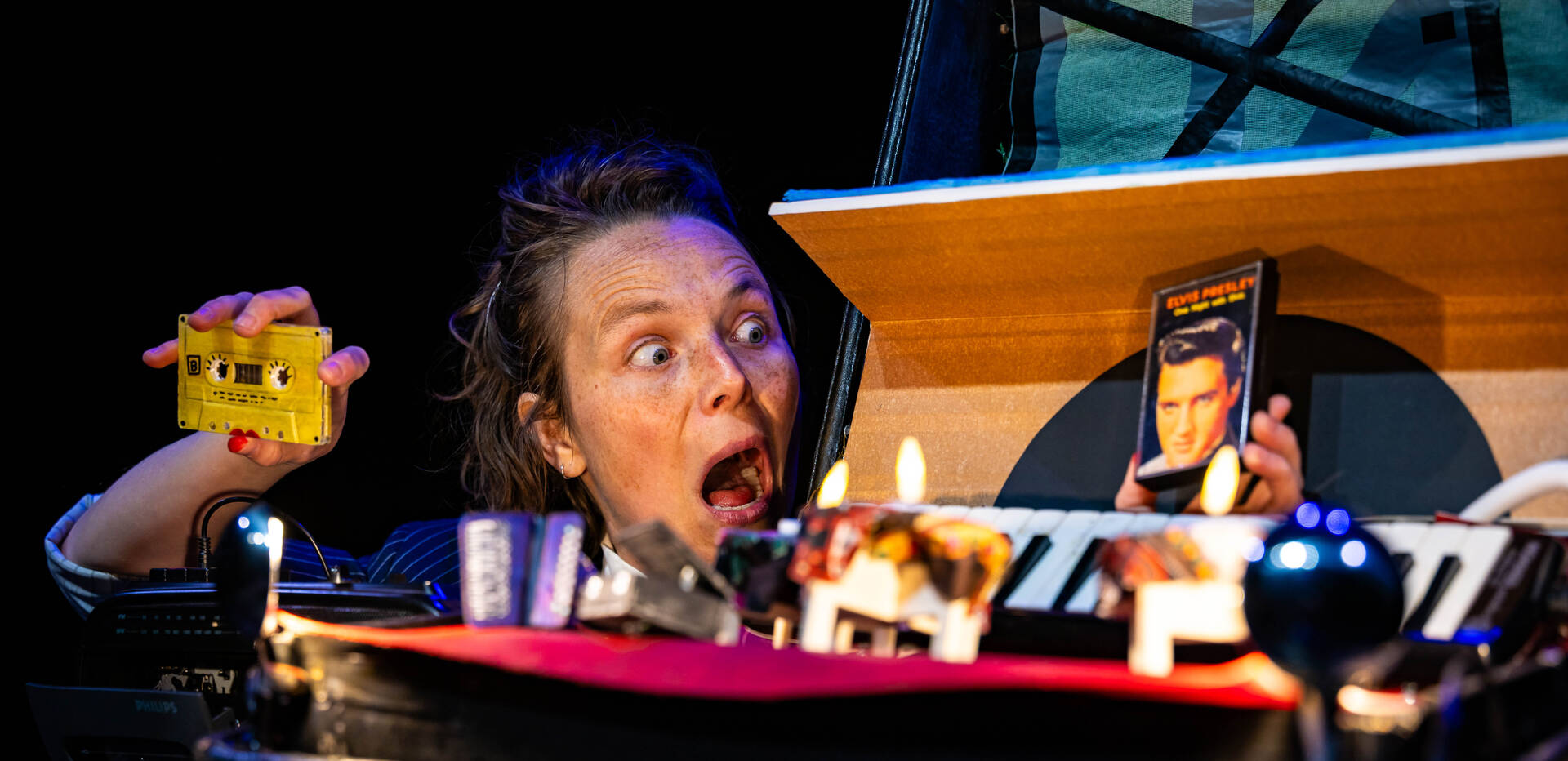 Actrice met verbaasde uitdrukking, houdend een gele speelgoedfiguur en een smartphone, met een tafel vol voorwerpen.