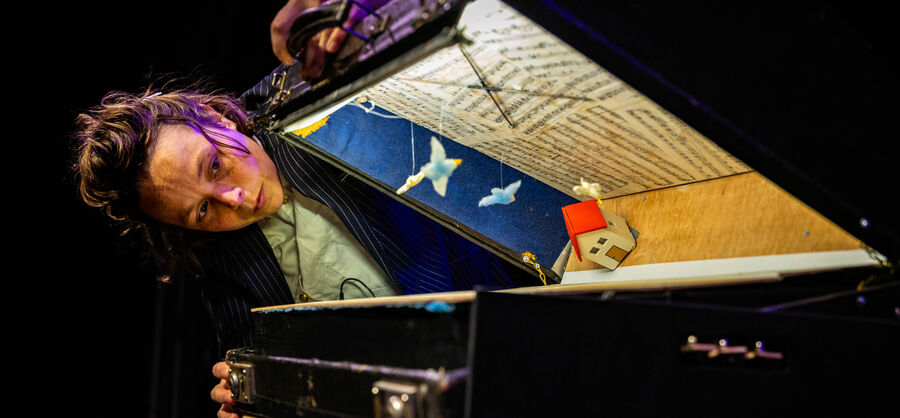 Een muzikant leunt over een open piano, terwijl hij een kleurrijke muziekdoos en noten bekijkt.
