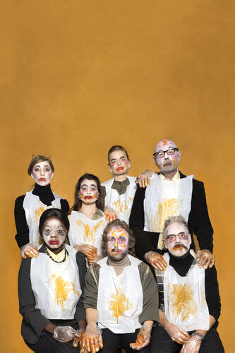 Groep mensen met kleurige gezichtsverf en schorten, poseren voor een oranje achtergrond.