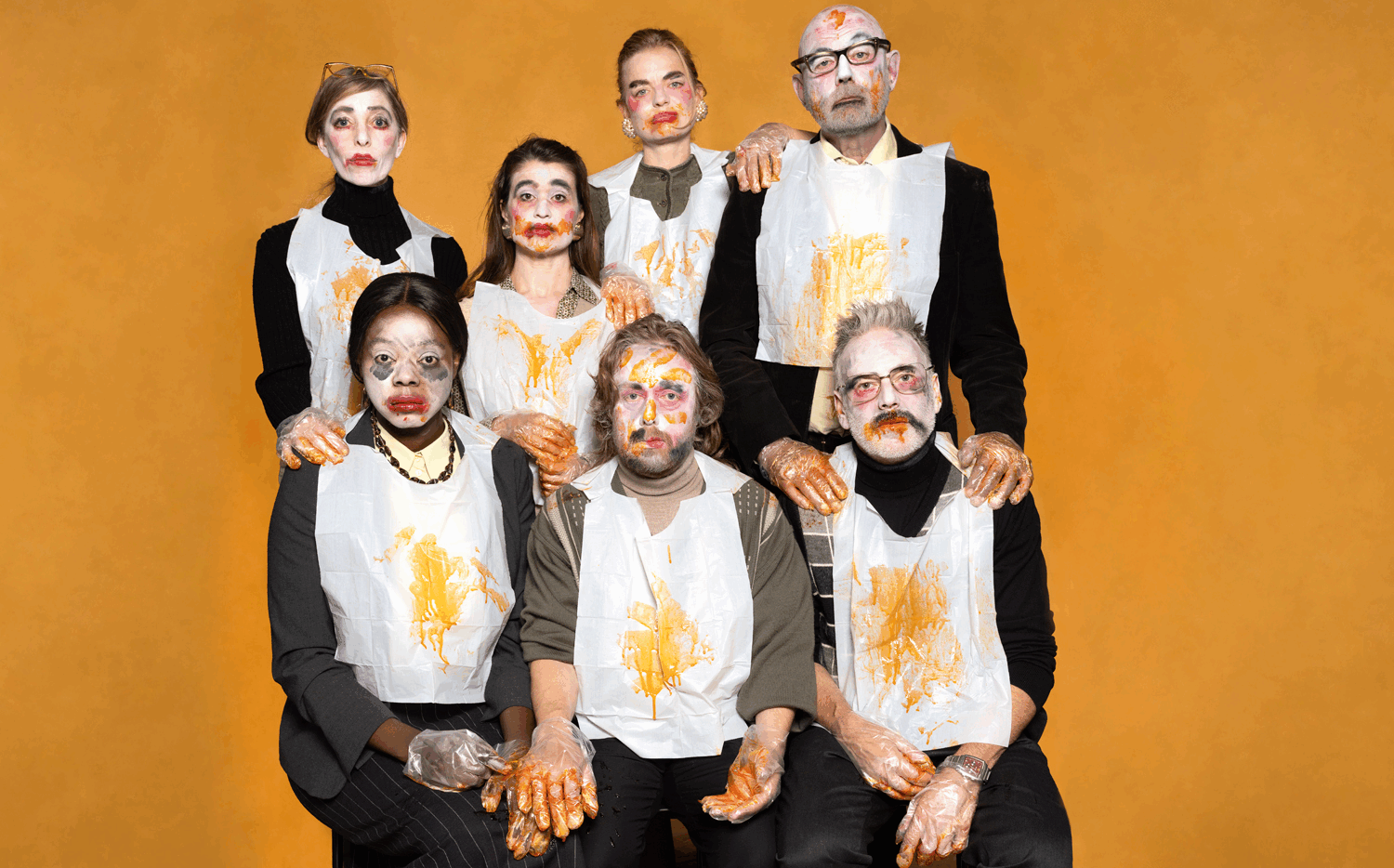 Groep mensen met geschilderde gezichten, gekleed in witte schorten met oranje patronen, tegen een oranje achtergrond.