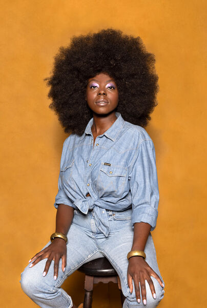 Vrouw met een grote afro-haarstijl, zittend op een kruk tegen een oranje achtergrond, gekleed in een denim outfit.