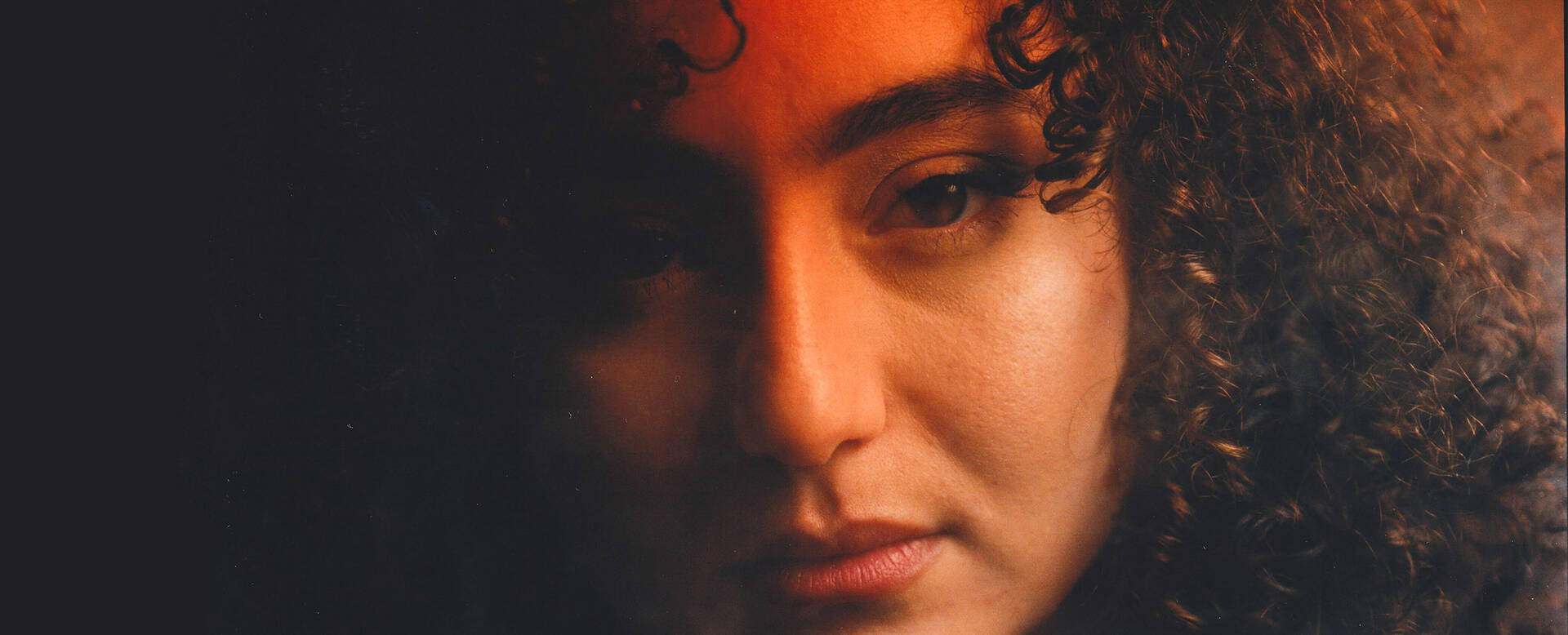 Een close-up van een vrouw met krullend haar, die een introspectieve uitstraling heeft in zachte verlichting.