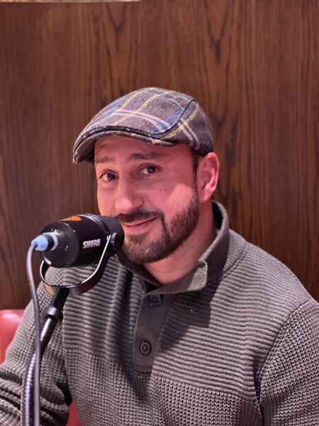 Man met een pet, zittend achter een microfoon in een warme, houten omgeving.