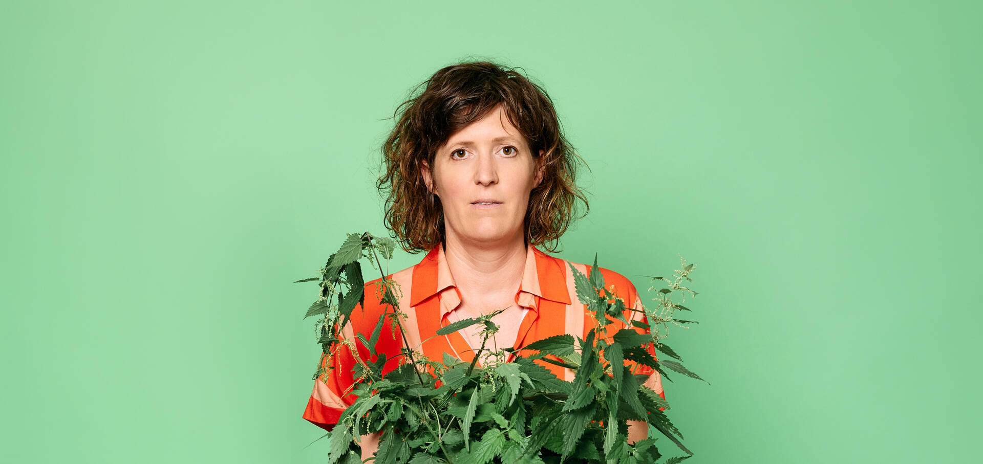  vrouw met krullend haar in een kleurige blouse houdt een plantenpot voor zich tegen een groene achtergrond.