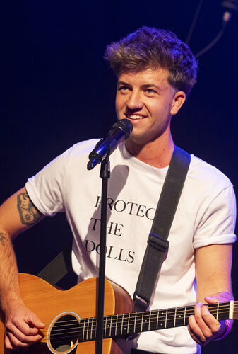 Zanger met gitaar op het podium, lachend en een T-shirt met de tekst "PROTECT THE PELLS".