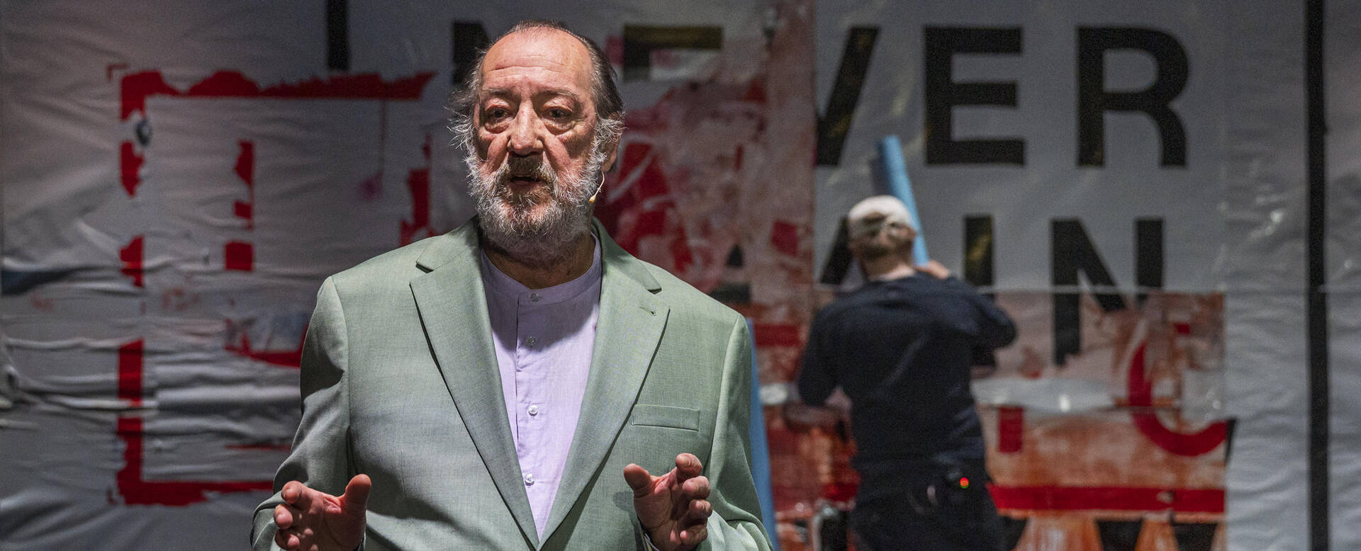 Een man in een lichtgroen kostuum staat voor een achtergrond met tekst en een tweede persoon aan het werk.