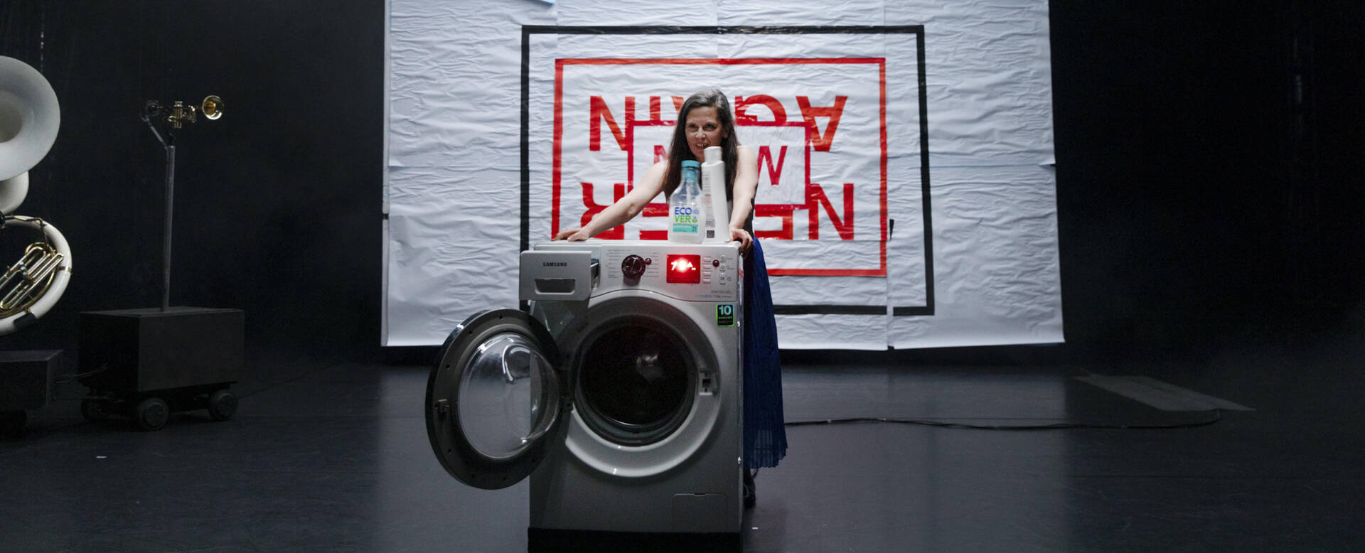 Een jonge vrouw staat naast een wasmachine, met een grote kunstinstallatie op de achtergrond.